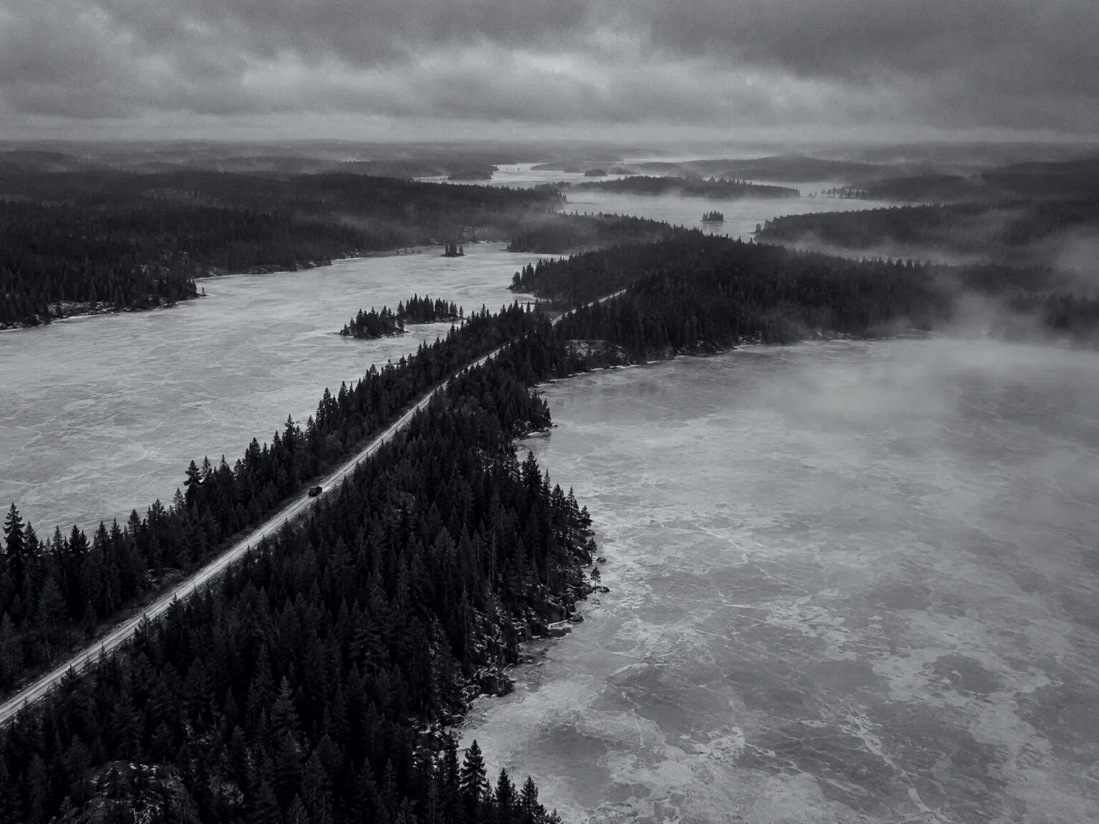Aerial view of a road on a strip of forest between two frozen lakes, one small vehicle on the road.